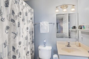 Une salle de bain comprenant des toilettes, un lavabo, un miroir, un porte-serviettes et un rideau de douche orné d’un motif de coquillages.