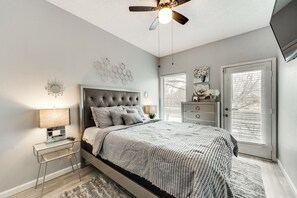 Une chambre à coucher avec une tête de lit capitonnée, un lit avec une literie à motifs, une table de chevet avec une lampe et un ventilateur de plafond.