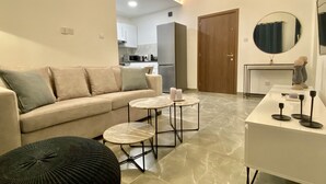 Un salón moderno con un sofá color beige, dos mesas redondas y un otoman negro. Se ve una puerta de madera y una cocina con armarios blancos al fondo.