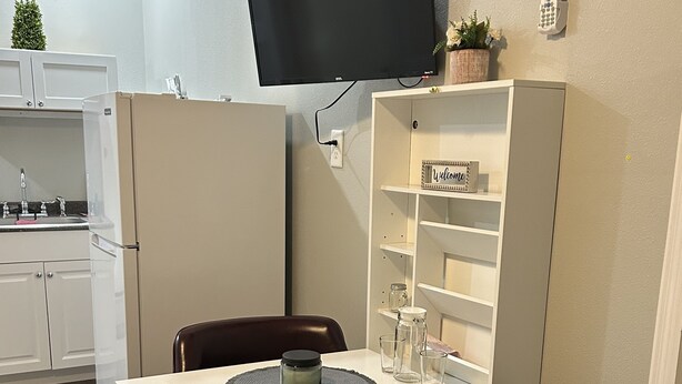 Une petite cuisine équipée comprenant un réfrigérateur, un évier et un petit plan de travail. Un t éléviseur est fixé au mur, au-dessus d’une armoire blanche sur laquelle est apposée une pancarte « Bienvenue ».