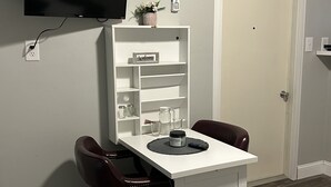 Une petite pièce, bien rangée, avec un bureau, deux chaises, un téléviseur fixé au mur, une porte blanche et une étagère sur laquelle est posé un panneau indiquant « Bienvenue ».