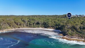 An aerial view of a coastal area with a beach, dense forest, and a clear sky.