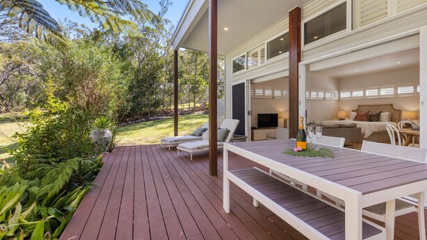 Una terraza con asientos al aire libre, una mesa con una botella y vasos, y vista a un dormitorio con cama y televisor.