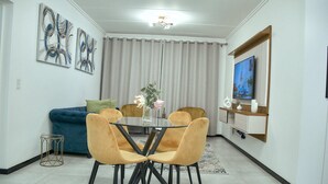 A dining area with a glass table, four chairs, a blue sofa, and a TV on the wall. - Medon Luxury Suites with power back up (Sandton)