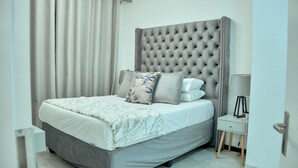 A modern bedroom with a tufted headboard, a grey bedspread, and a bedside table with a lamp. - Medon Luxury Suites with power back up (Sandton)