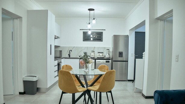 A modern kitchen with white cabinets, a stainless steel refrigerator, and a dining area with a glass table and yellow chairs. - Medon Luxury Suites with power back up (Sandton)
