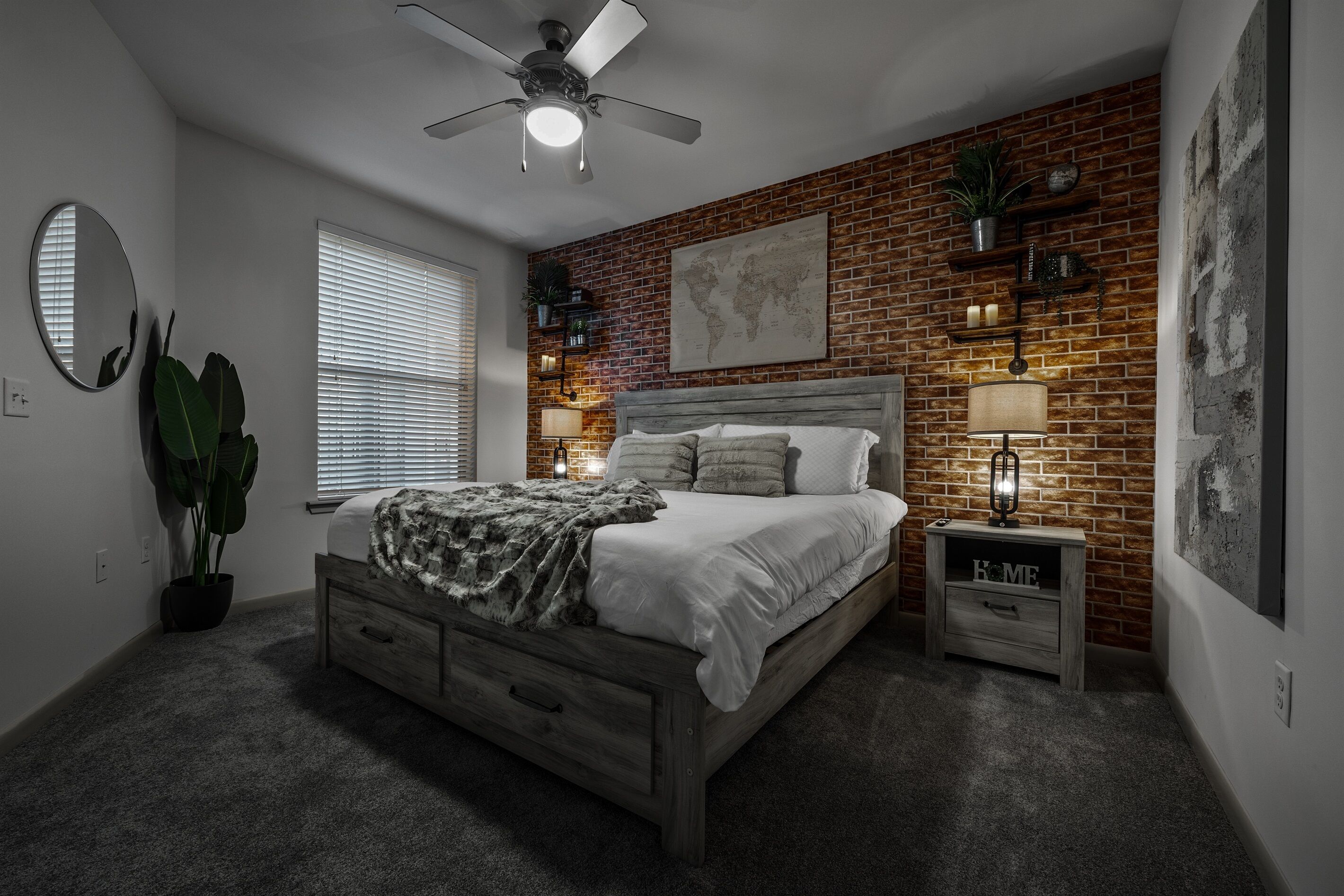 Un dormitorio con pared de ladrillo, cama con cabecera de madera, mesita de noche con lámpara, ventilador de techo y una ventana grande con persianas.