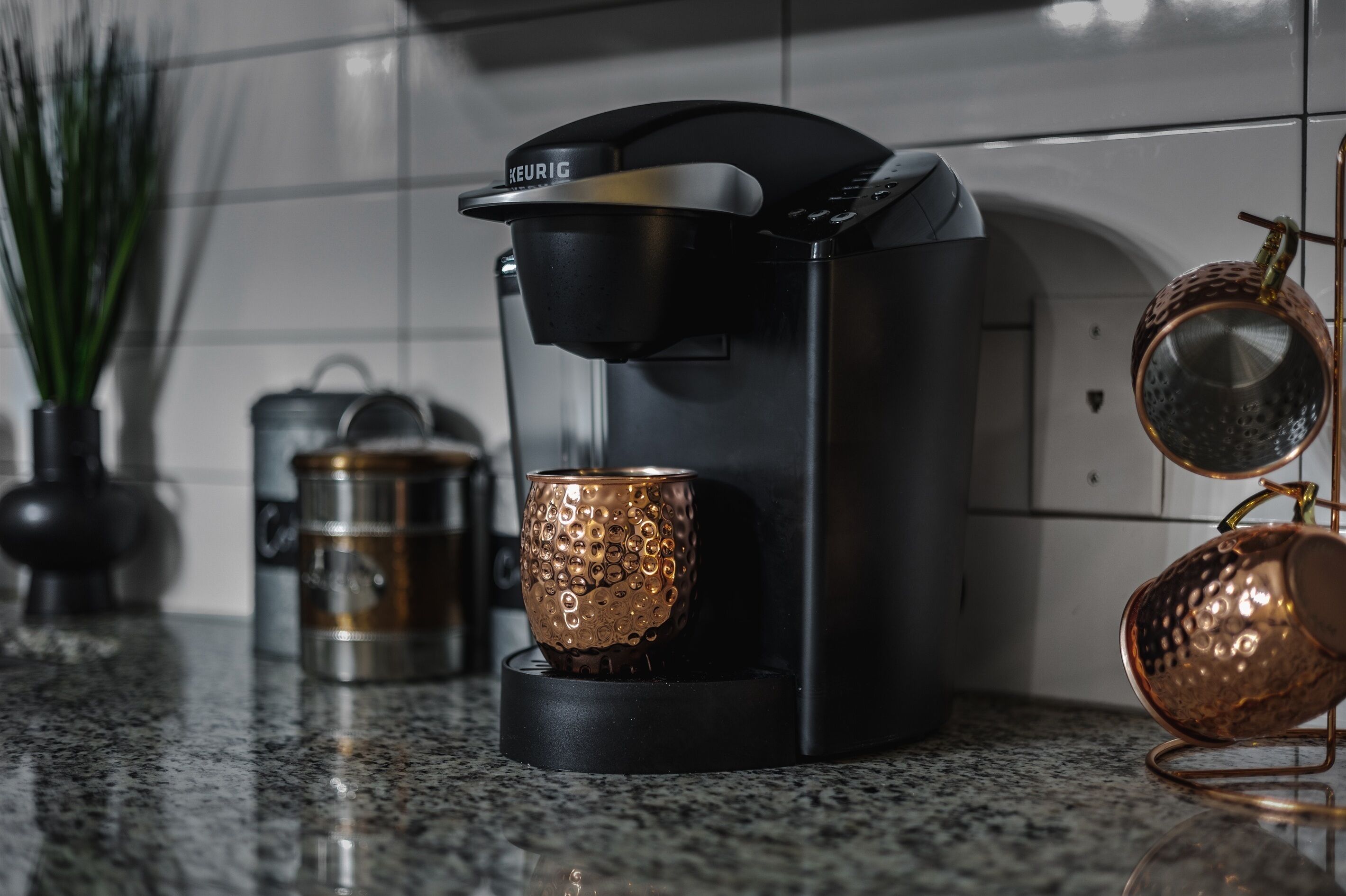 A modern kitchen with a black coffee maker, a stainless steel kettle, and two copper mugs on a granite countertop.