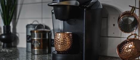 A modern kitchen with a black coffee maker, a stainless steel kettle, and two copper mugs on a granite countertop.