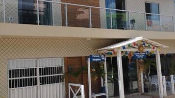 Un edificio de varios pisos con un área de entrada cubierta, un acceso con portón y un pequeño espacio de descanso con sillas y mesas.