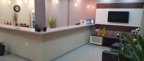 Zona de recepción con mostrador, plantas en macetas, reloj, televisor y un área de descanso con cojines.