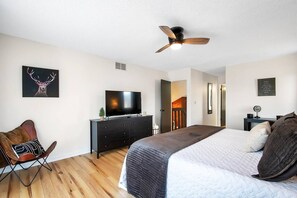 A bedroom with a bed, a TV on a dresser, a ceiling fan, and a door leading to another room. - Trade chilly Kansas nights for a soak in the private hot tub, then rally the crew for 300+ arcade games. Your fall basecamp in northwest Wichita. (Wichita)