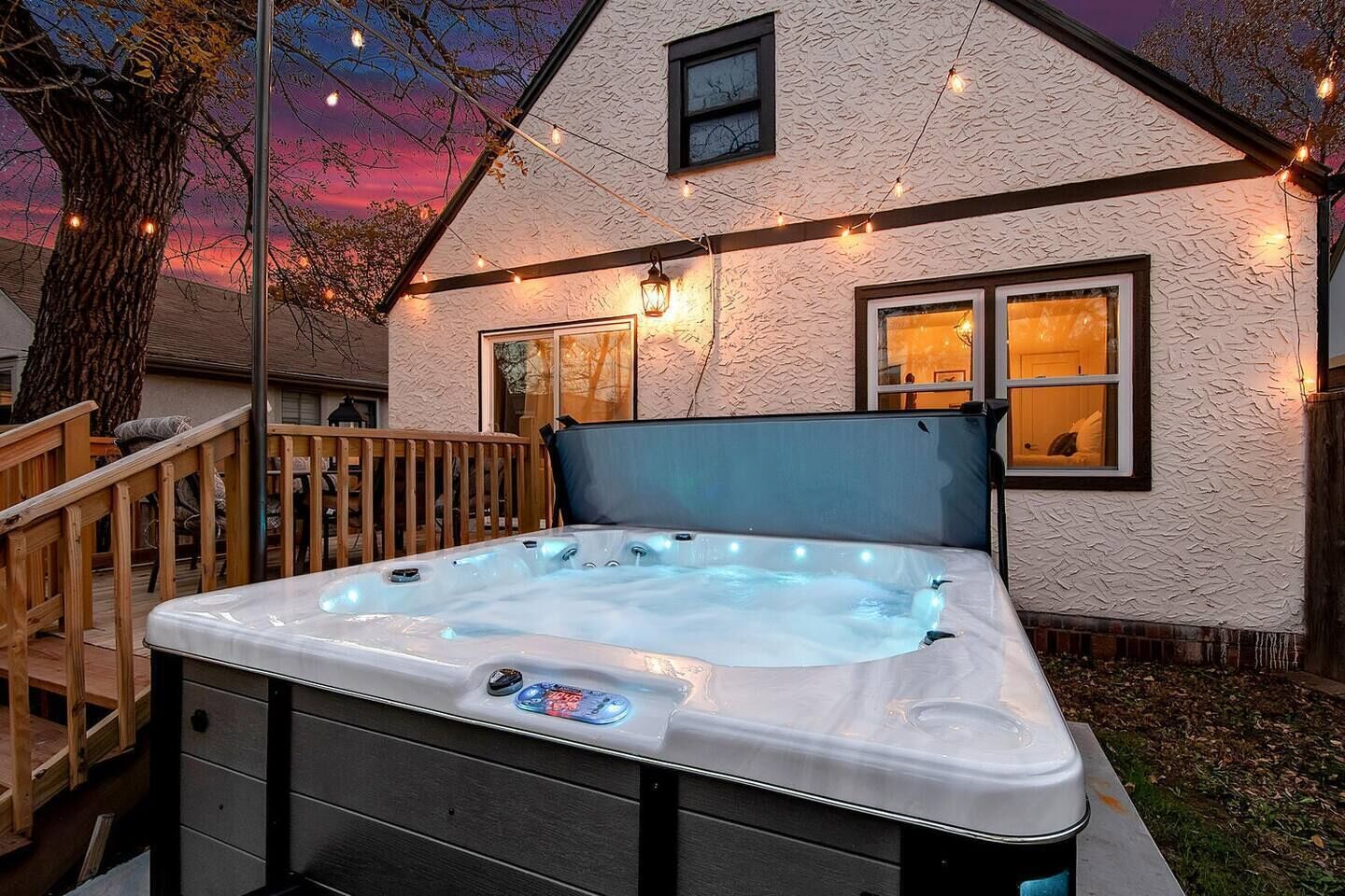 A hot tub in a backyard with a wooden deck, a tree, and string lights.