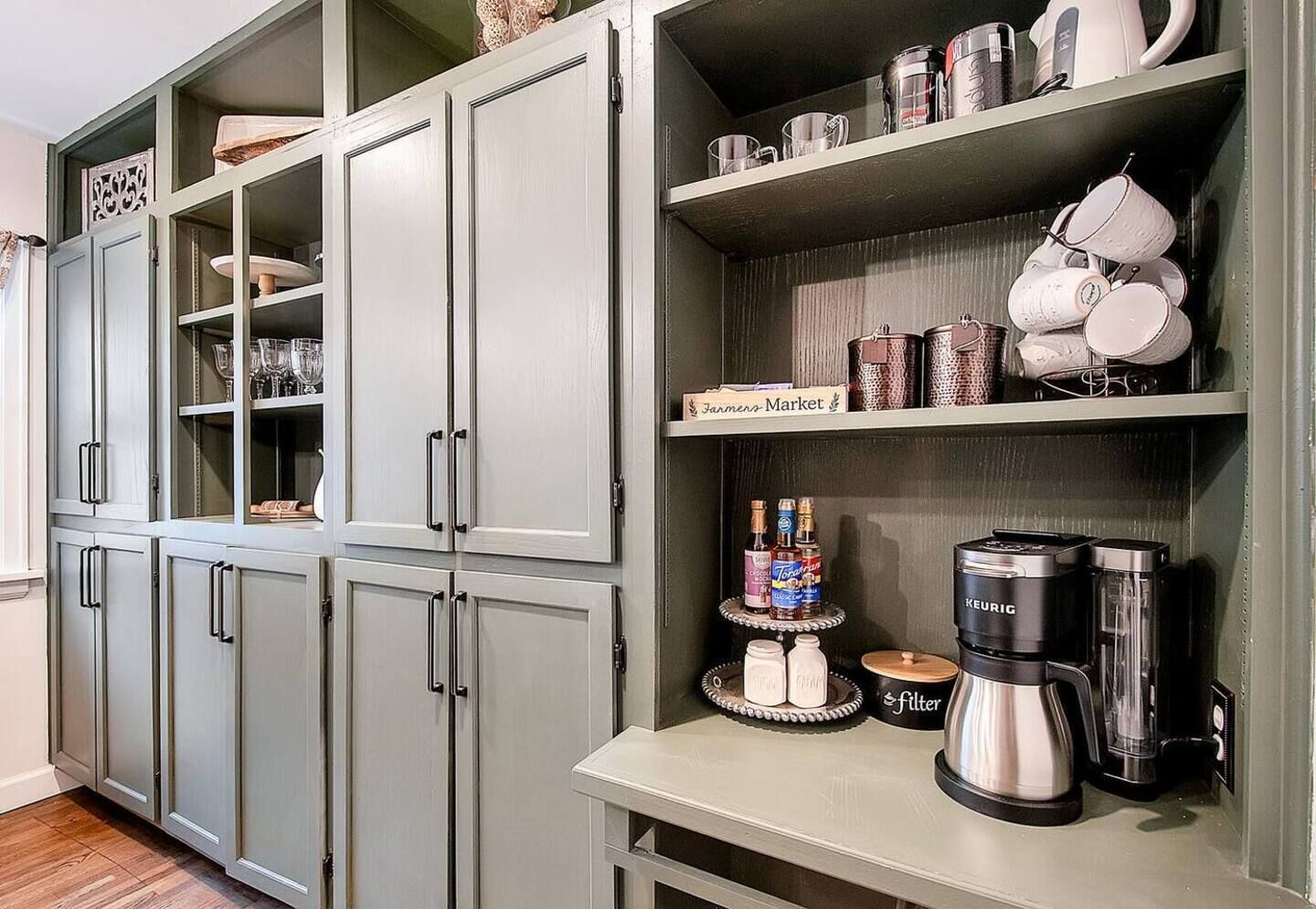 A kitchen with a built-in coffee maker, a kettle, and a shelf with mugs and containers.