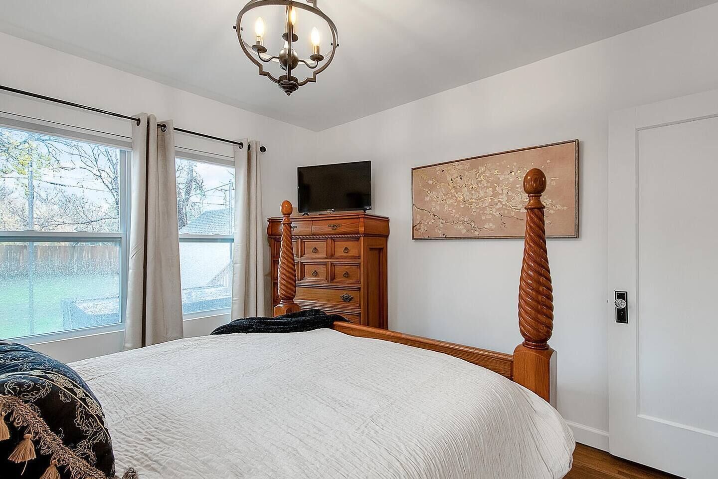 A bedroom with a large bed, a wooden dresser, a television on the wall, and a window with curtains.