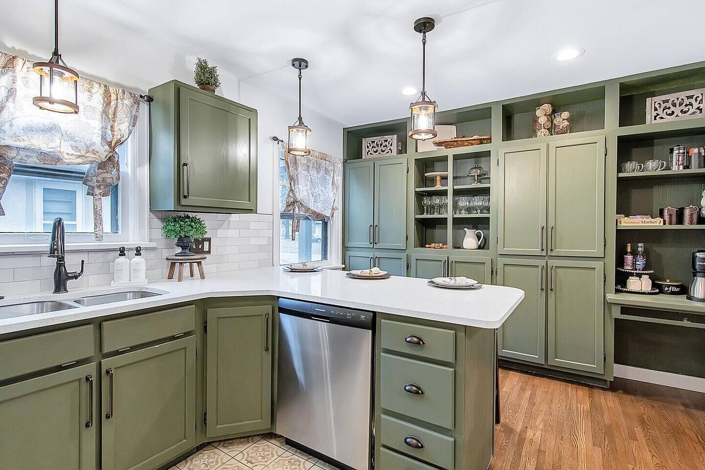 A kitchen with green cabinets, a white countertop, and a dishwasher. There are hanging pendant lights and a window with a view.