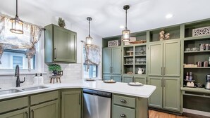 A kitchen with green cabinets, a white countertop, and a dishwasher. There are hanging pendant lights and a window with a view. - Riverside Renaissance - Spa!  King Beds! Beautiful Renaissance decor (Wichita)