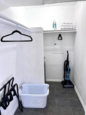 A laundry room with a white wall, a hanging clothes rack, a white laundry basket, a vacuum cleaner, and a small shelf with folded towels.