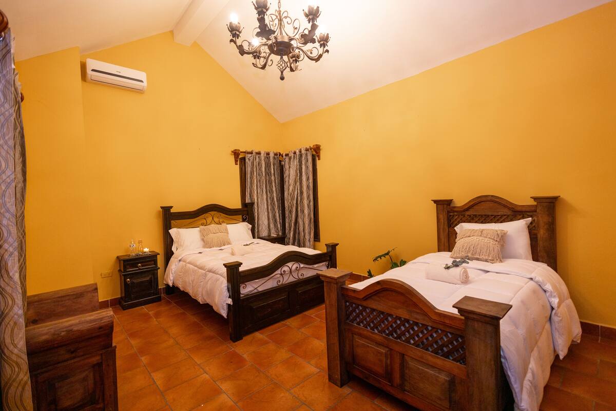 Une chambre avec deux lits en bois, un lustre, un appareil de climatisation et une petite table de chevet avec une lampe.