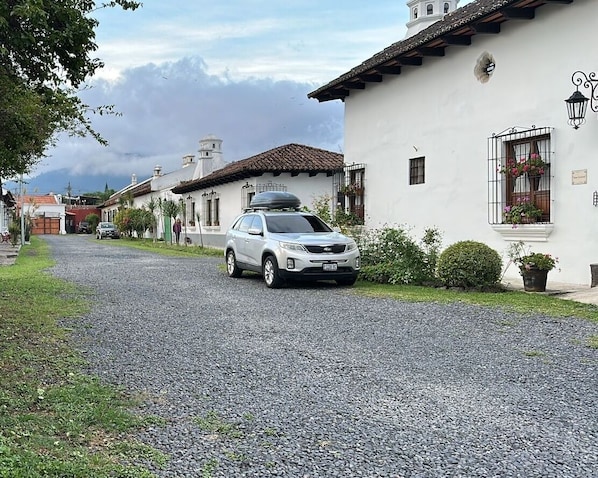 A gravel road leading to a white building with a tiled roof, a parked car, and a small garden area.