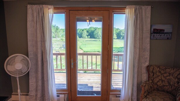 Una habitación con puerta de madera, un ventilador, una silla y vista a un área con césped a través de la puerta.