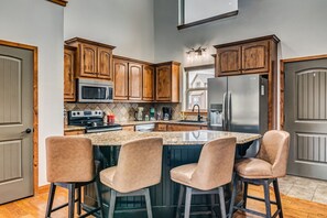 A kitchen with wooden cabinets, a stainless steel refrigerator, a microwave, and a stove. There are three bar stools and a large island with a granite countertop.