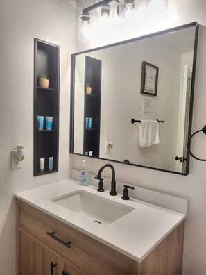 A bathroom with a white sink, black faucet, and a large mirror above it. There are shelves with toiletries and a potted plant.