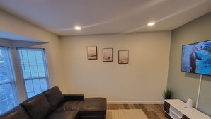 A modern living room with a sectional sofa, a coffee table, and a TV mounted on the wall. There are three framed pictures on the wall and a small plant on the side table. - Near University of Delaware, Christiana Care Hospital, & the Christiana Mall! (Newark)