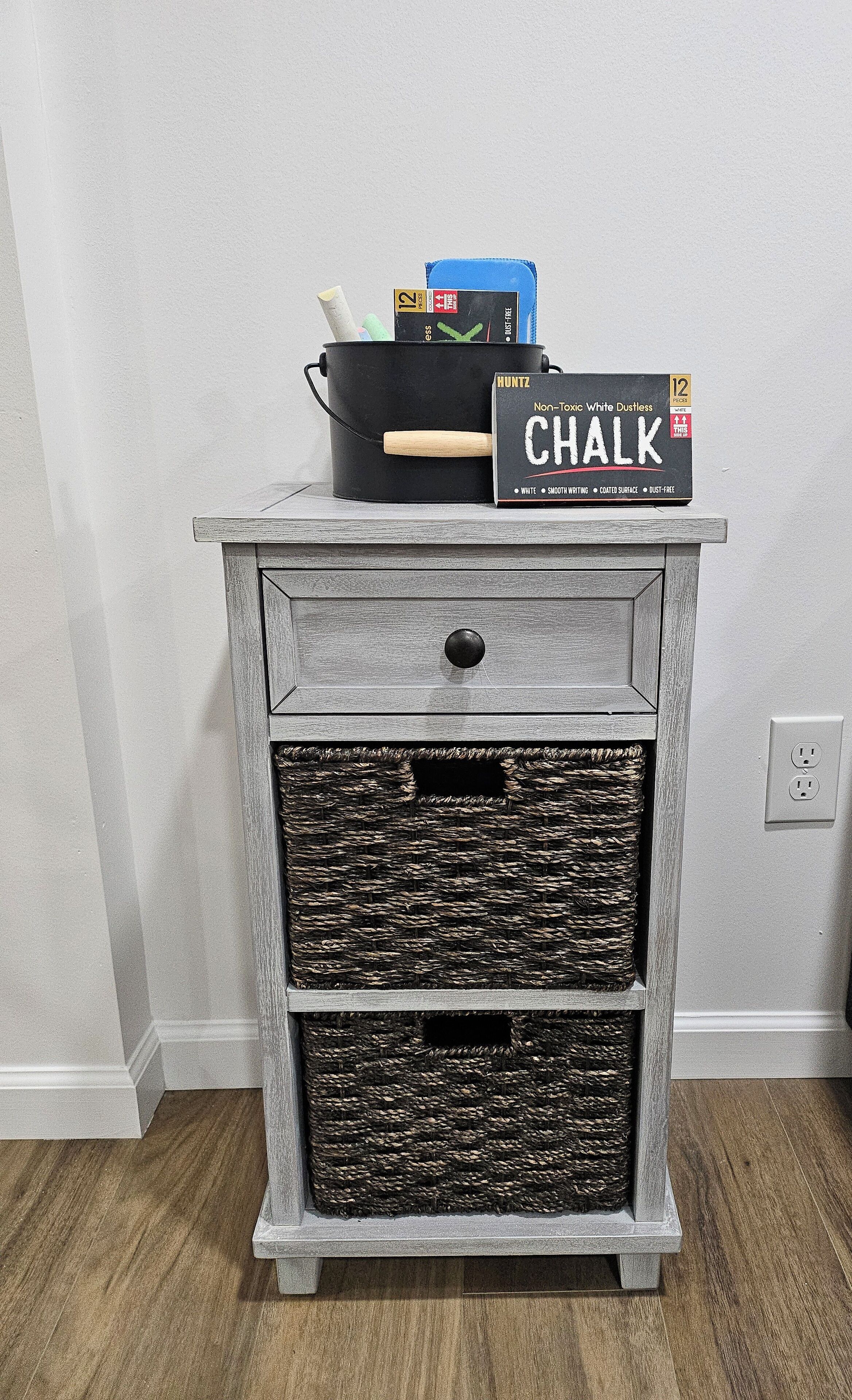A rustic wooden nightstand with a chalkboard sign, a woven basket, and a black pot with writing utensils.