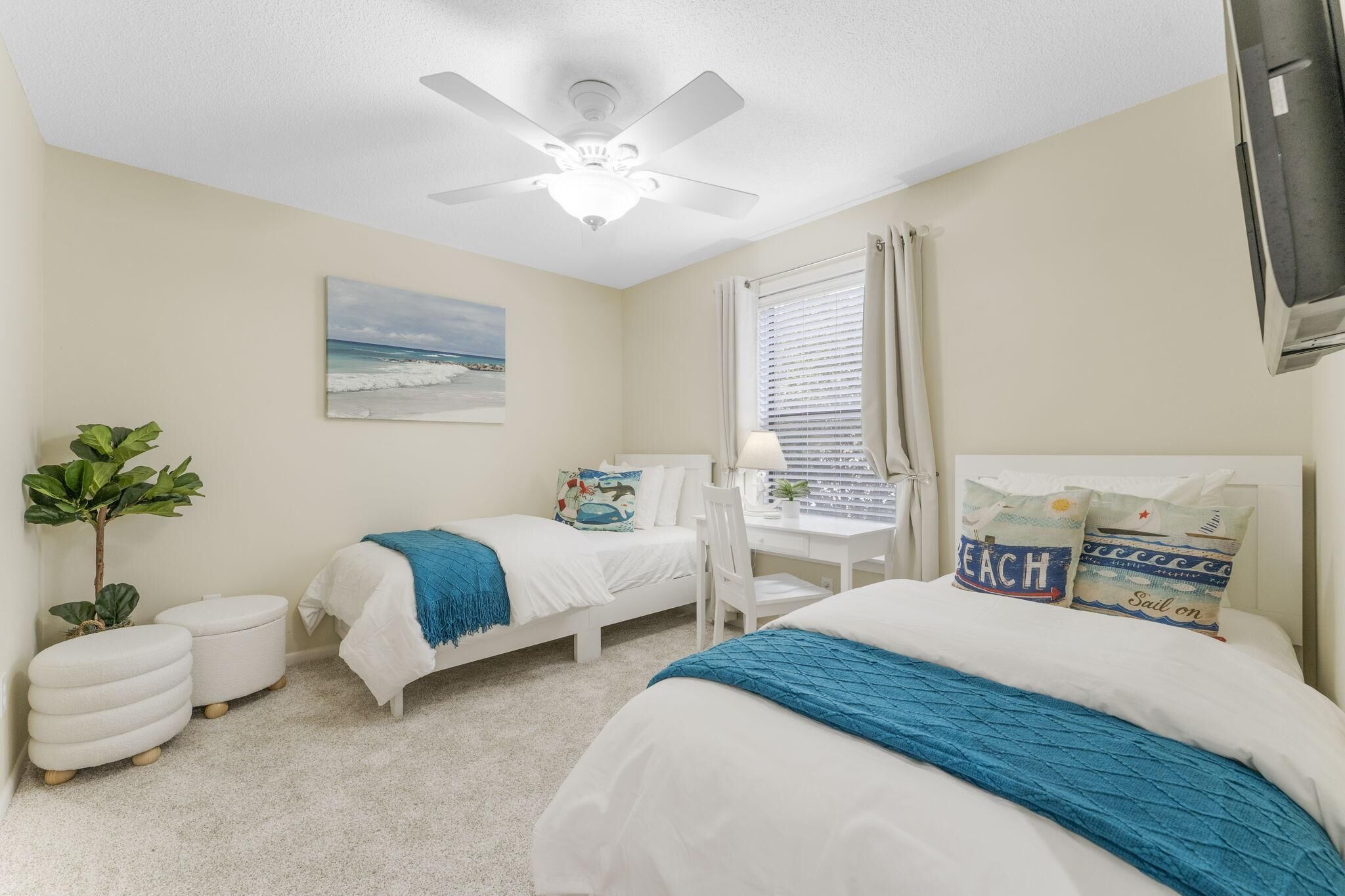 A bedroom with two beds, a ceiling fan, a desk with a chair, a window with blinds, and a plant.