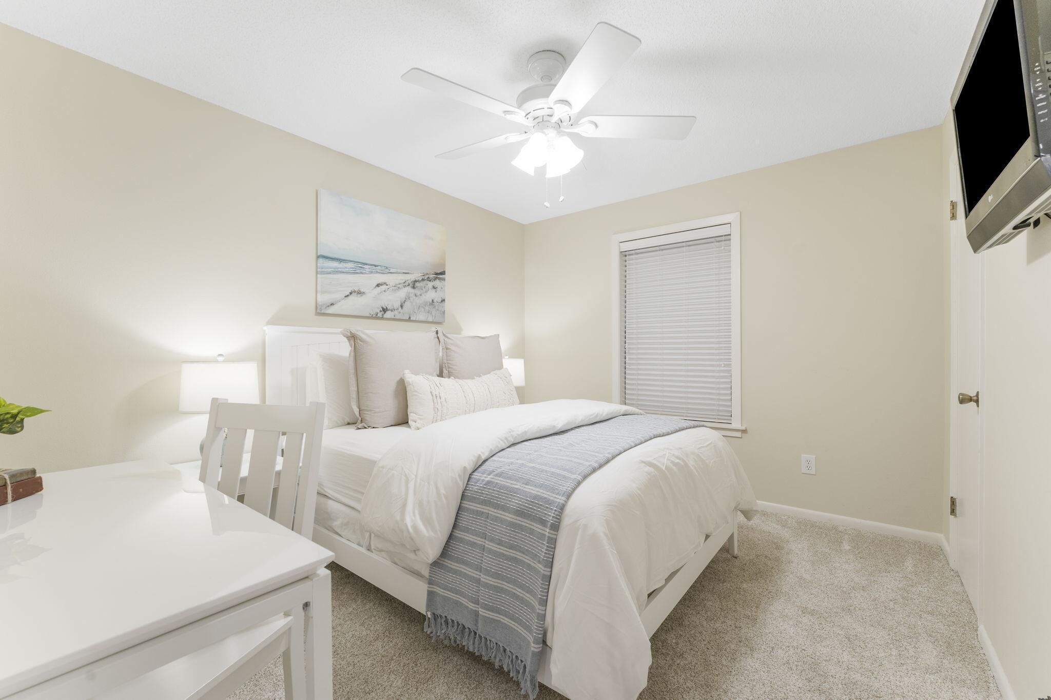 A bedroom with a bed, a desk, a chair, a ceiling fan, a window with blinds, and a TV mounted on the wall.