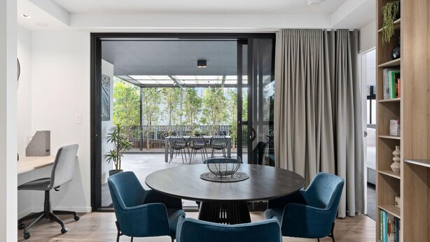 Un comedor moderno con una mesa redonda y cuatro sillas, una estantería y vistas a un balcón con barandilla y vegetación.