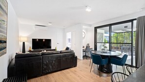 Una sala moderna con un sofá de cuero negro, un televisor de pantalla plana y un comedor con sillas azules y una mesa redonda.