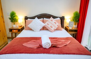 Un lit bien fait, recouvert d’une couverture rouge, avec des oreillers blancs et une serviette blanche enroulée. Il y a deux tables de chevet avec des lampes et des plantes en pot.