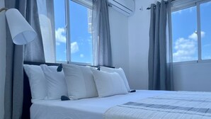 Un llit ben fet amb llençols blancs i una manta estampada, una làmpada de nit i una vista del cel a través d'una finestra amb cortines.