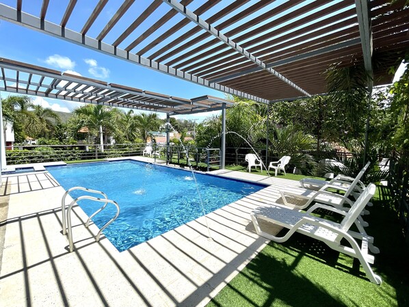 A swimming pool with a wooden deck and white lounge chairs, surrounded by greenery and a pergola structure.