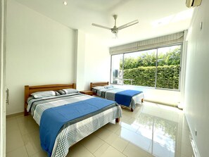 A hotel room with two beds, a ceiling fan, an air conditioning unit, and a large window with a view of greenery.