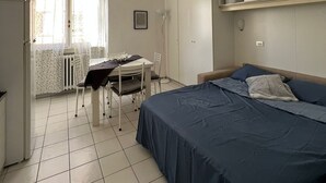 Une petite pièce propre, avec un lit, une table, des chaises et une fenêtre avec des rideaux.