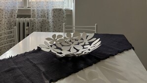 Une pièce avec une fenêtre ornée de rideaux à motifs floraux, un radiateur, une table blanche sur laquelle se trouve un vase décoratif, un tissu noir et un tableau encadré accroché au mur.