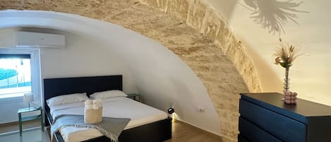 A bedroom with a curved stone wall, a bed, a dresser, and a small table with a lamp.