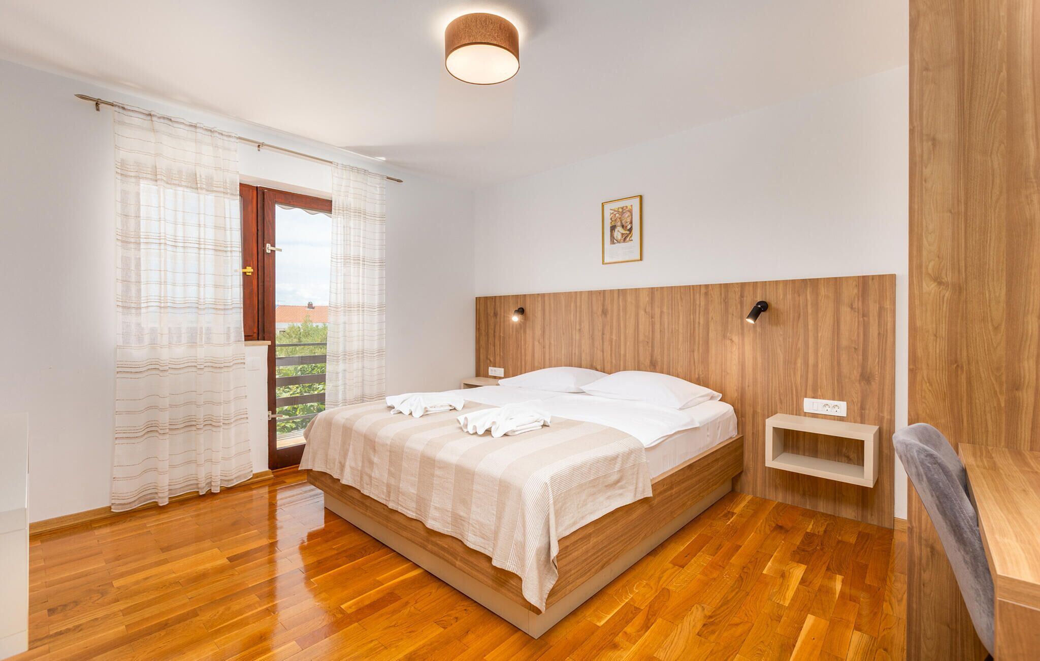 Un dormitorio con cabecera de madera, cama con ropa blanca, una mesita de noche y una ventana con cortinas.