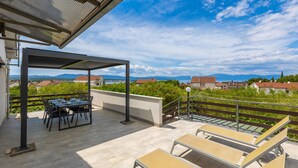 A covered outdoor patio with a table and chairs, a sun lounger, and a view of a mountainous landscape.