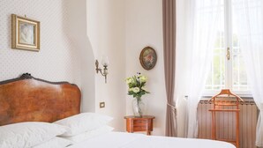 En pænt redt seng med hvide sengetøj, et træhovedgærde, et lille natbord med en vase blomster, og et vindue med lette gardiner.