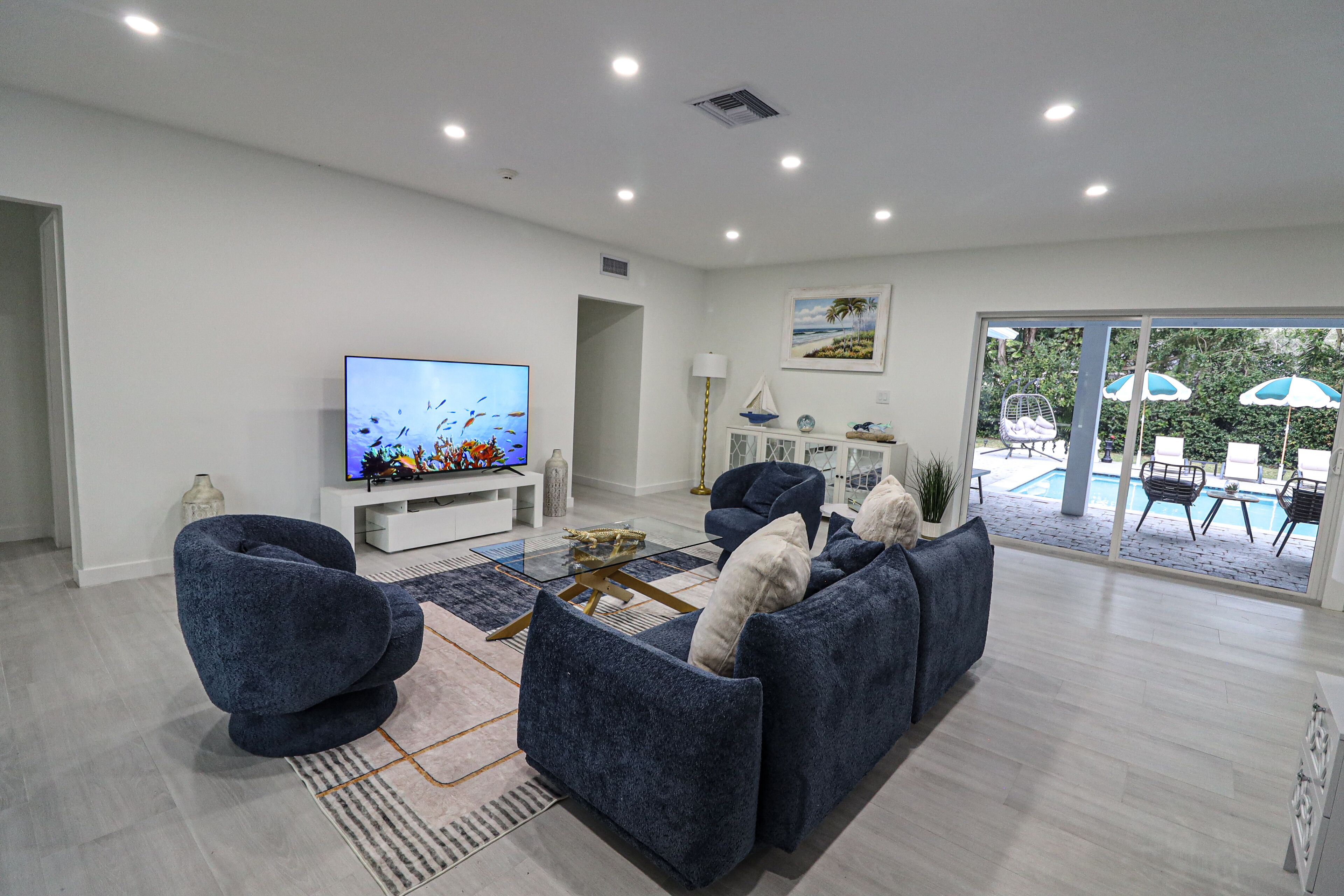 A modern living room with a flat-screen TV, blue sofas, a coffee table, and a view of an outdoor pool area.