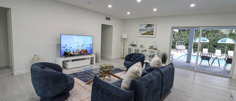 A modern living room with a flat-screen TV, blue sofas, a coffee table, and a view of an outdoor pool area.