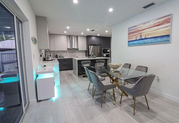 A modern kitchen with white cabinets, a central island, and a dining area with a glass table and grey upholstered chairs. - Villa Cristina in Miami (Miami)