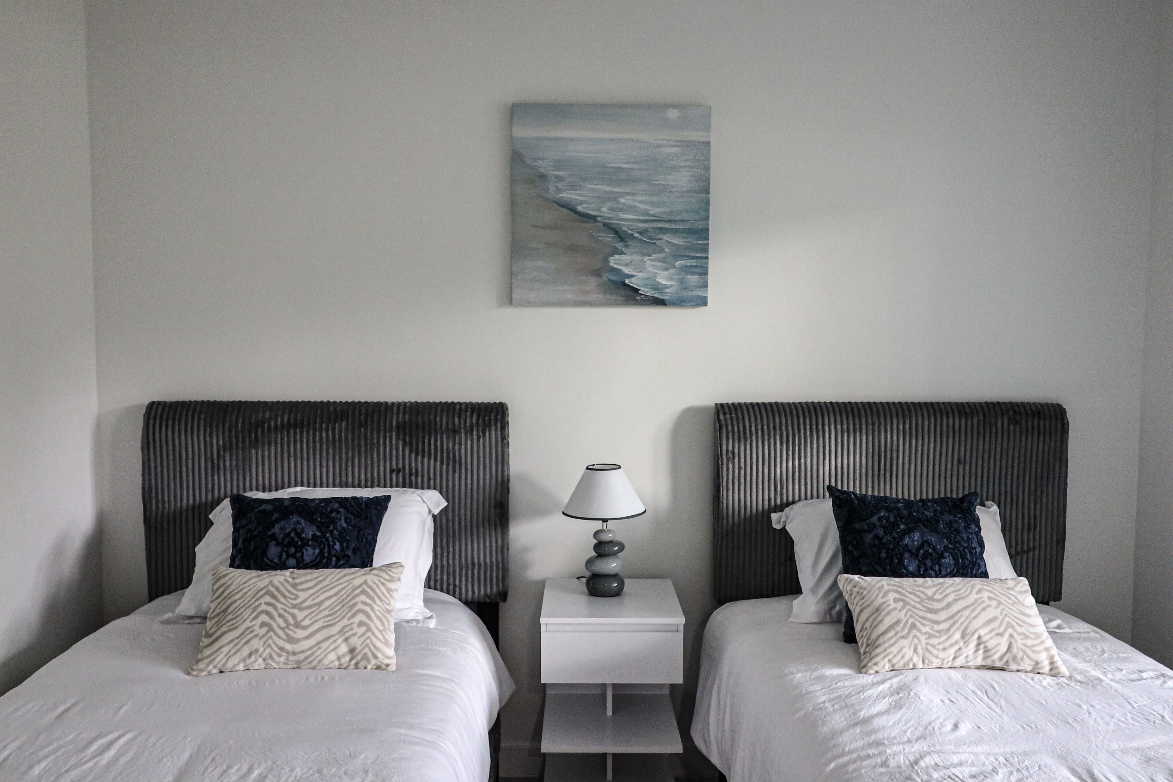 Une chambre d’hôtel avec deux lits, chacun doté d’une tête de lit, d’une table de chevet avec une lampe et d’un tableau représentant une scène de plage au mur.