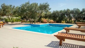 Et rektangulært pool med fliseterrasse, omgivet af træloungestole og en række træer.