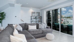 Ein modernes Wohnzimmer mit einem grauen Ecksofa, einem runden Pouf und einem schwarzen Regal mit Dekoration.