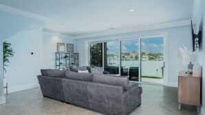 Ein geräumiges Wohnzimmer mit grauem Sofa, einem Holzschrank und einer großen Schiebeglastür, die auf einen Balkon mit Blick auf das Wasser führt.